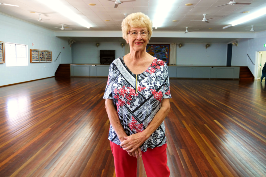 a woman stands smiling at the camera with her hands folded, inside a community hall