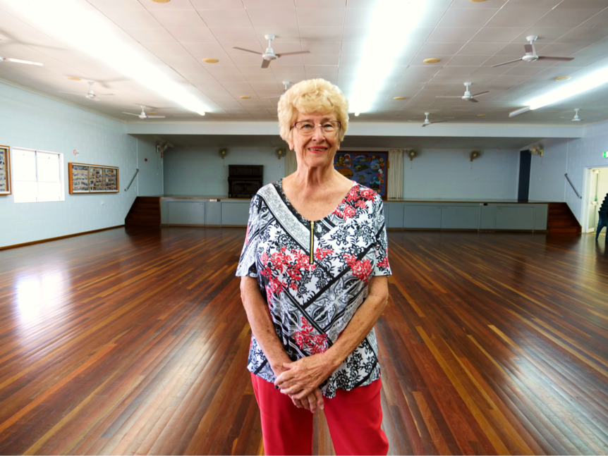 a woman stands smiling at the camera with her hands folded, inside a community hall