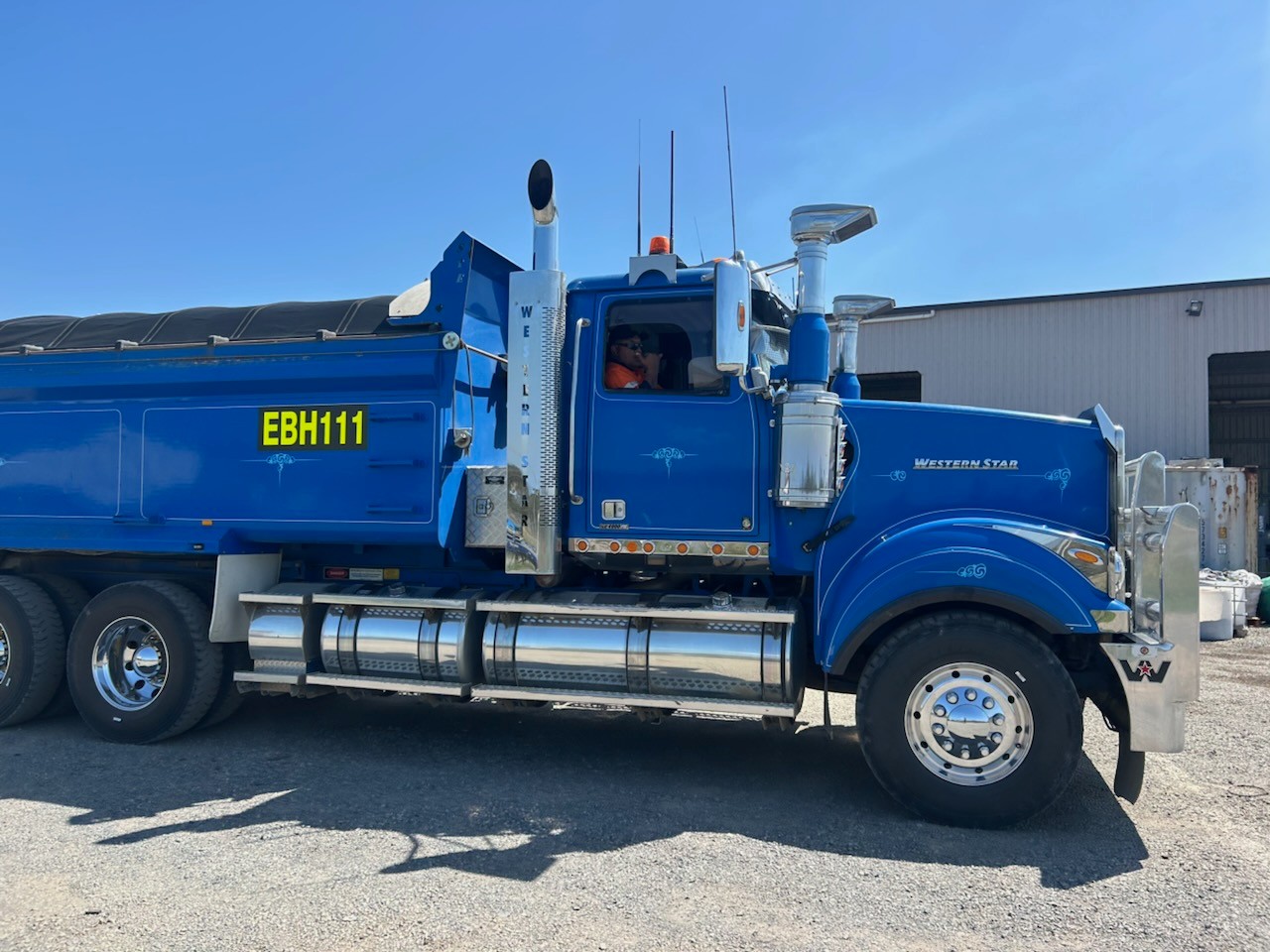 Una toma larga de un gran camión azul con un Johnnie Tumai con una camiseta naranja sentado en el asiento del conductor.