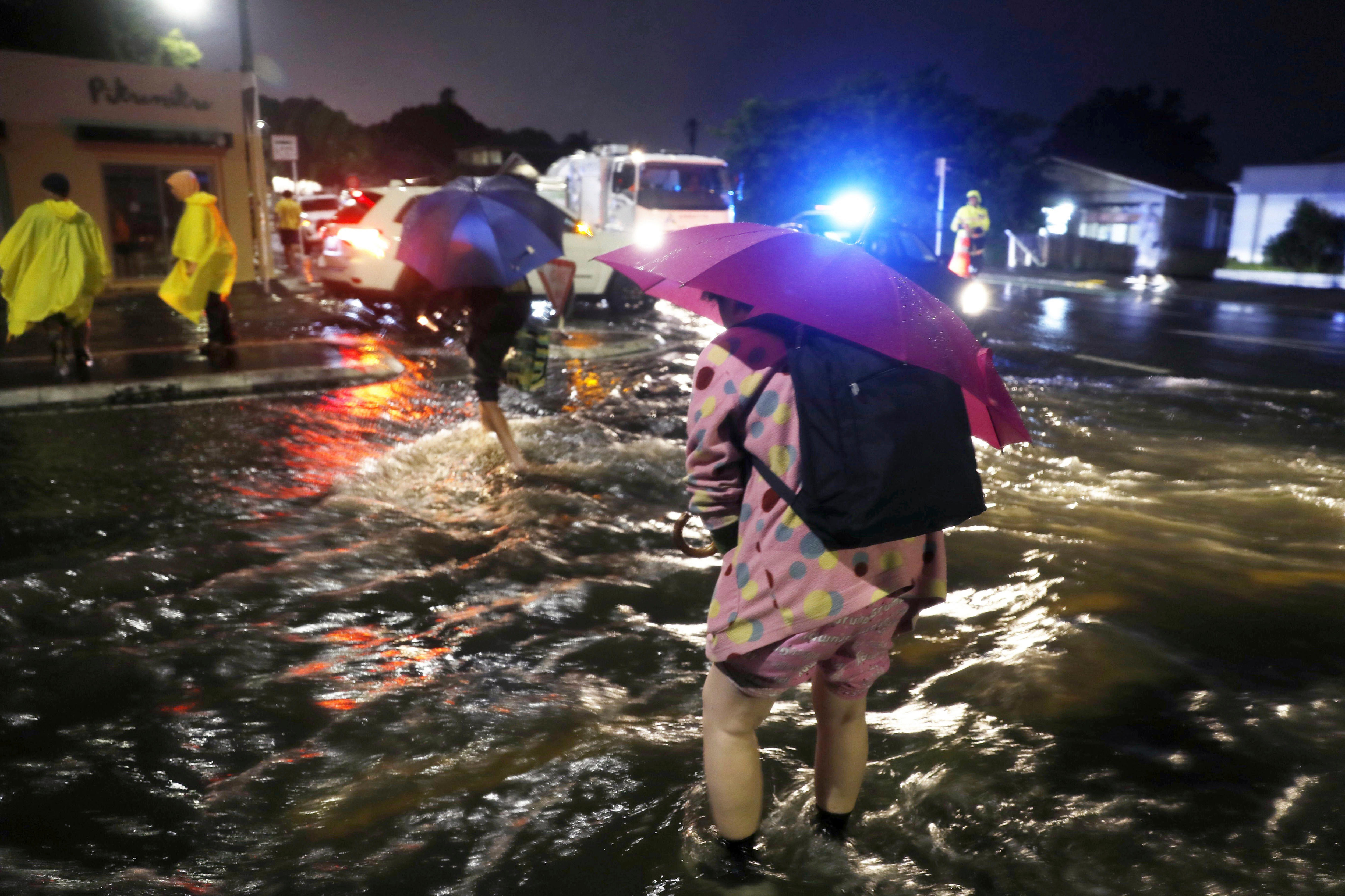 带着雨伞和雨衣的人们穿过被洪水淹没的街道晚上有齐脚踝的水。” class=