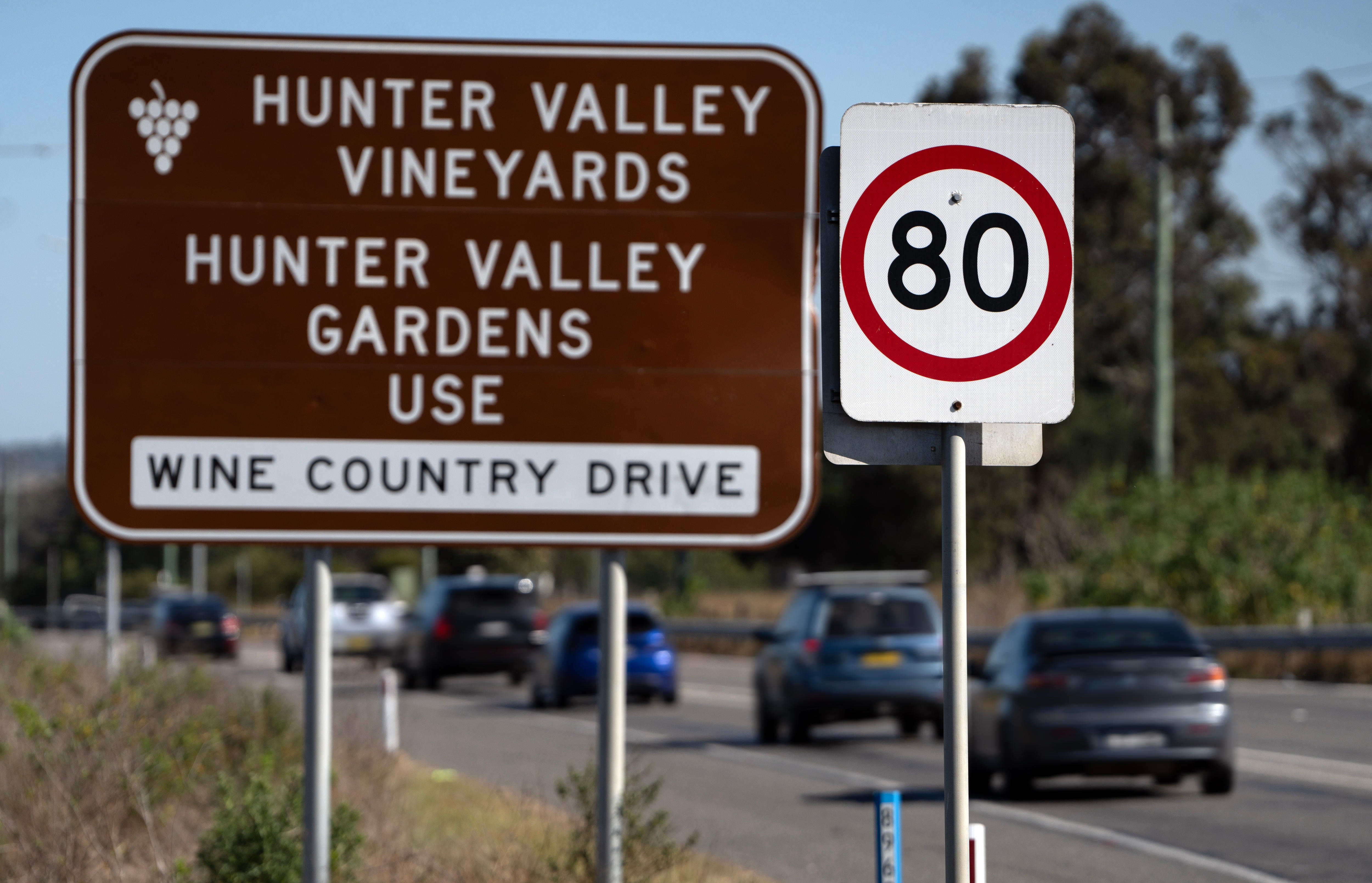 Multiple cars drive on a road past a brown sign reads 'Hunter Valley Vineyards...Wine Country Drive'. 80 speed sign in front.