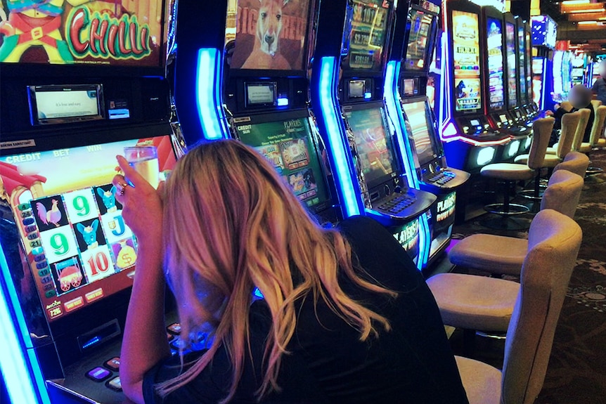 A blond woman sits with a drink at a pokie.