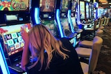 A blond woman sits with a drink at a poker machine.