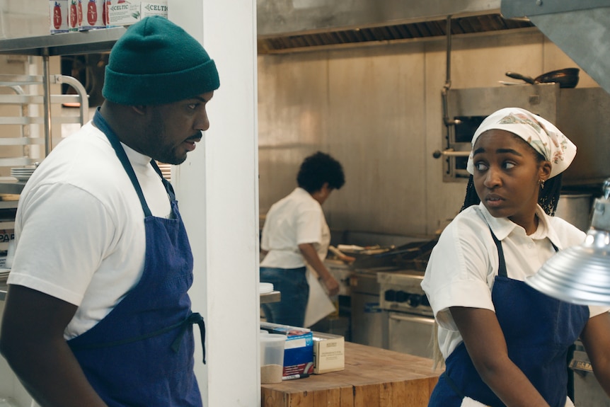 A young Black man and woman in white tshirts and blue aprons talk to each other in a commercial kitchen