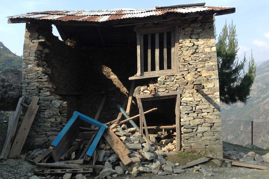 A badly damaged stone classroom with a tin roof and mountains in the background.