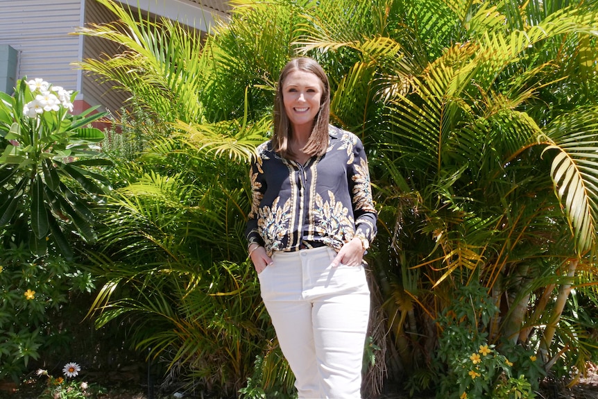 A woman in a black shirt and white pants stands infront of trees smiling at camera