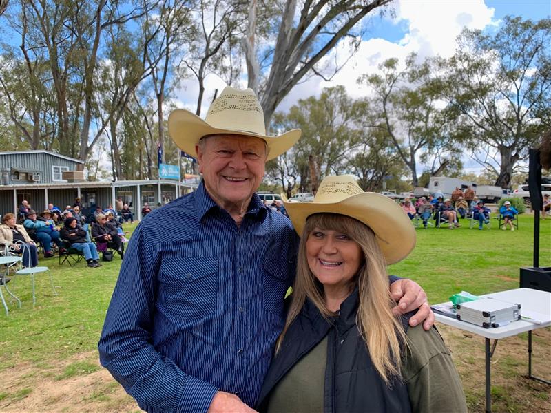 Man in blue long-sleeve shirt and cream akubra standing with his arm around a woman in a cream akubra and khaki shirt, smiling