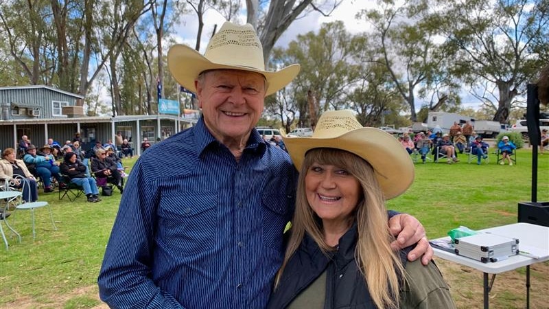 Man in blue long-sleeve shirt and cream akubra standing with his arm around a woman in a cream akubra and khaki shirt, smiling