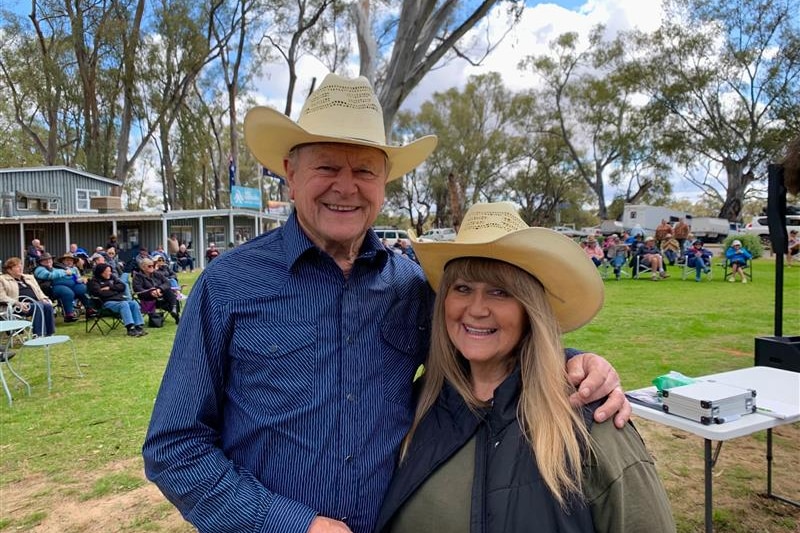 Man in blue long-sleeve shirt and cream akubra standing with his arm around a woman in a cream akubra and khaki shirt, smiling