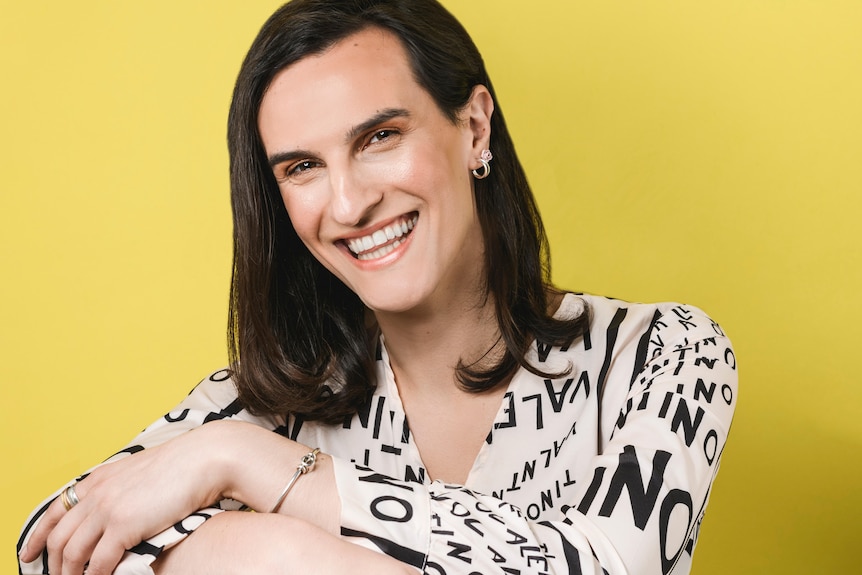 Bayley Turner, a trans woman, smiles brightly in front of a yellow background. She has her arms folded in front of her.