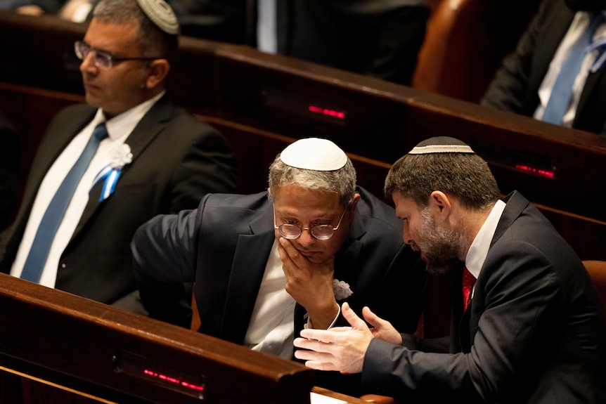 A man wearing a suit, sitting in parliament leans forward as another man talks to him.