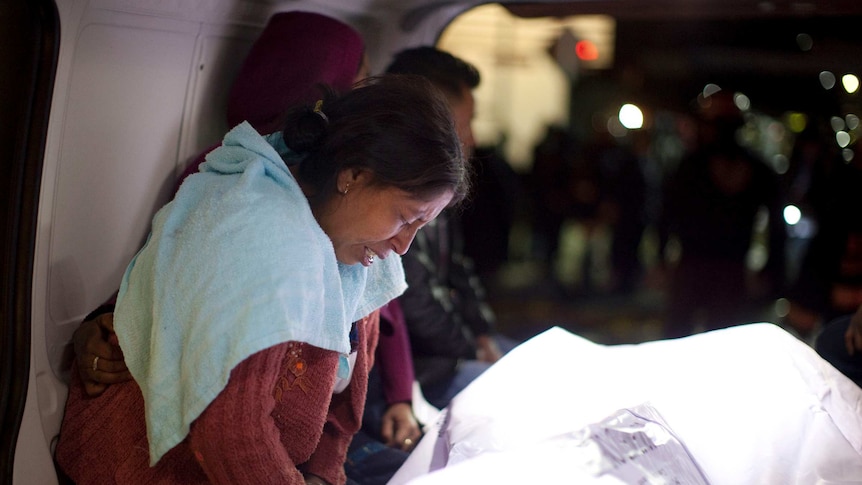 A women cries as she leans over a body, covered in a white sheet, in what appears to be the back of a van.