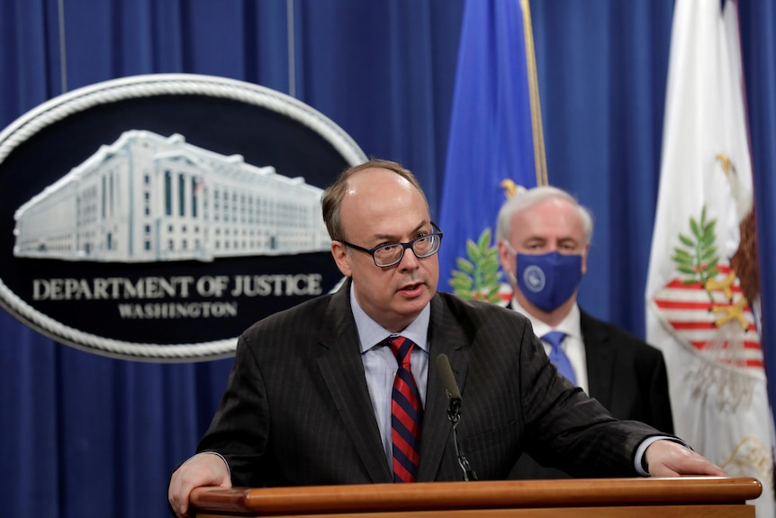 A man in a suit and black rimmed glasses stands at a lectern in front of a 'Department of Justice" sign
