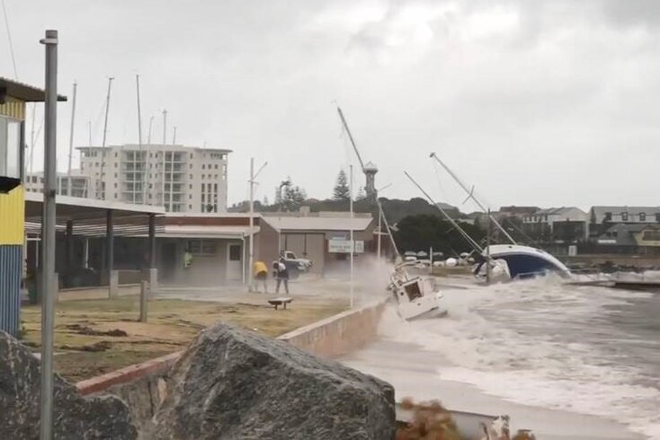 Loose yachts wash up against a concrete barrier at the Bunbury Yacht Club, while two people brace against the spray of water.