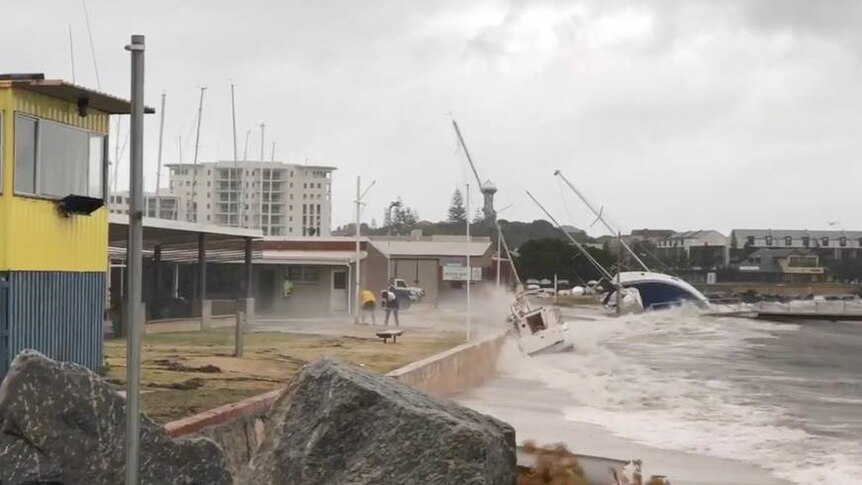 Loose yachts wash up against a concrete barrier at the Bunbury Yacht Club, while two people brace against the spray of water.