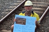 A protester wearing a hard hat and high vis holding a sign about species extinction sits on train tracks at Bowen Hills.