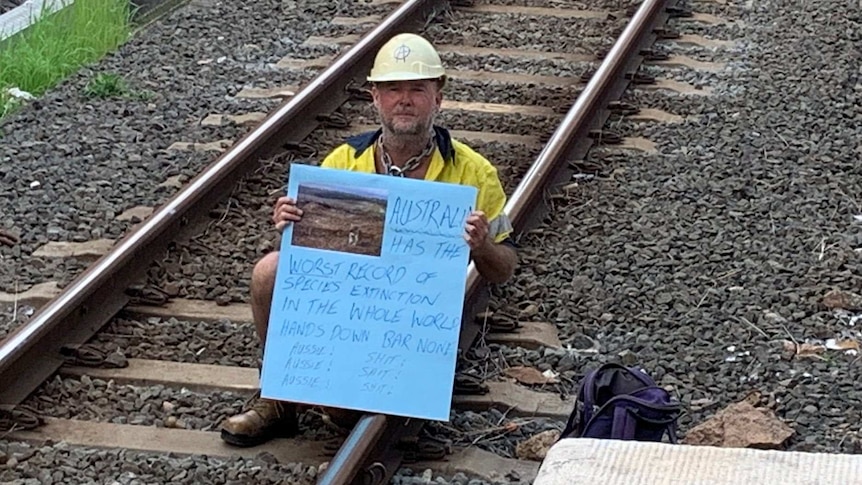 A protester wearing a hard hat and high vis holding a sign about species extinction sits on train tracks at Bowen Hills.