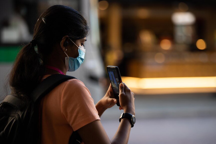 Girl with mask on and lon gblack hair takes a picture of a building with her phone.