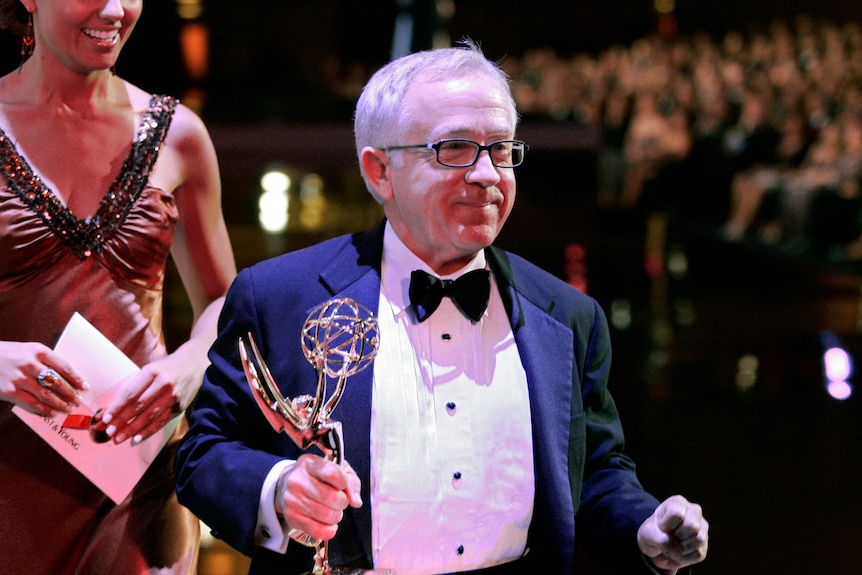 A man holds an Emmy in his right hand and looks off to the right. He is wearing a black and white tux and glasses. 