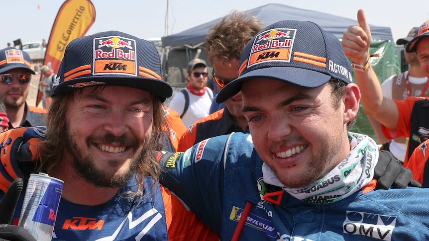 Two men stand smiling with their arms around each other wearing caps and motorcycle jackets