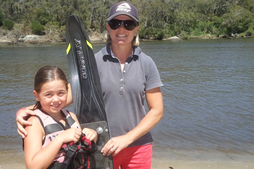 A mother with her arm around her young daughter, who's wearing a life jacket, and holding a water ski