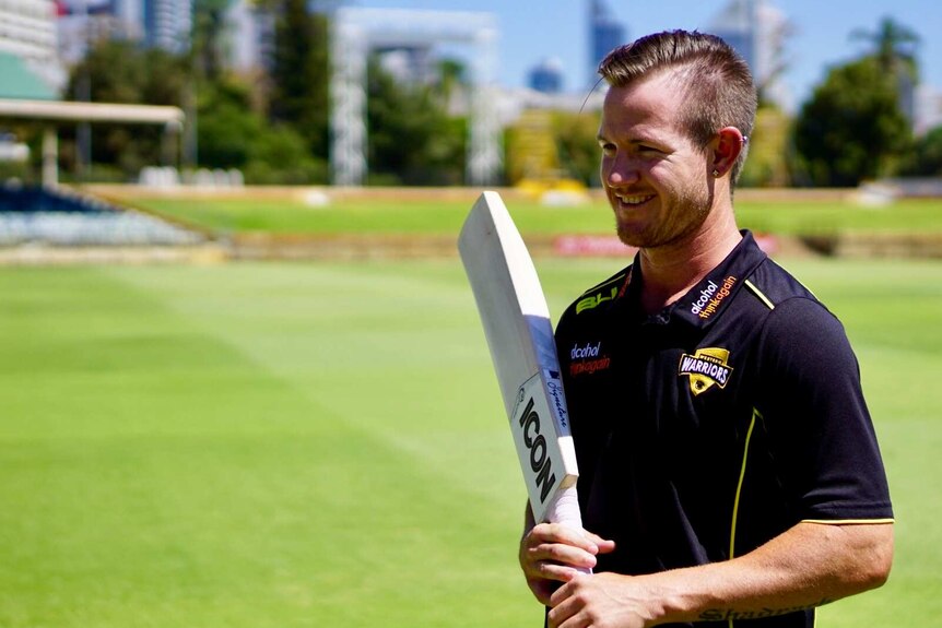 A cricket player wearing a polo shirt smiles and looks off-camera while holding a bat on a pitch.