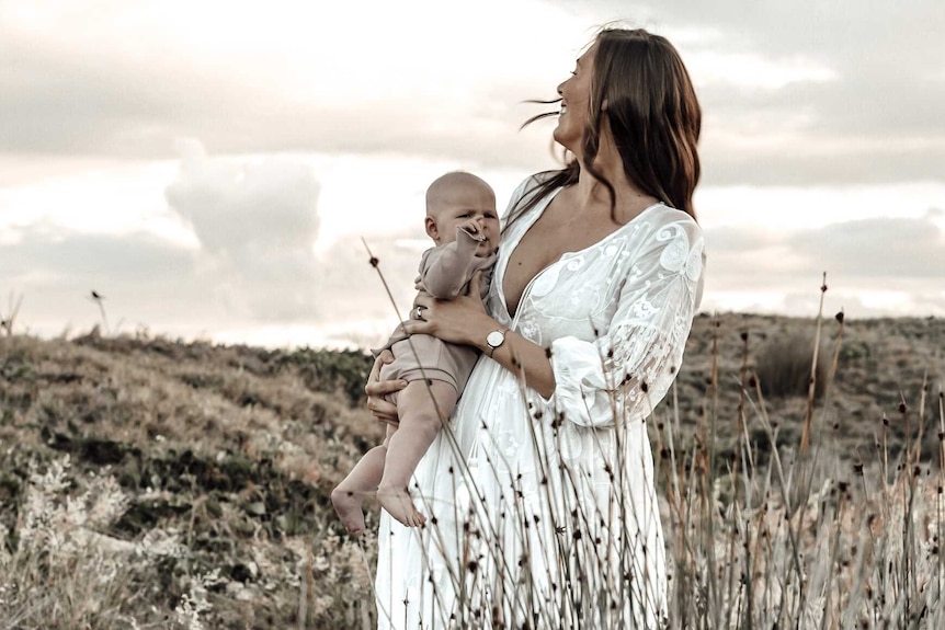 A mother in a loose white dress with long brown hair holds a baby wearing a white romper in a field.