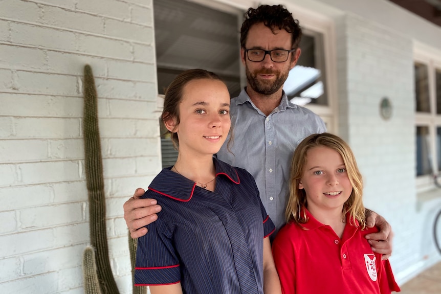 A man with his arm around his two smiling children out the front of their house.