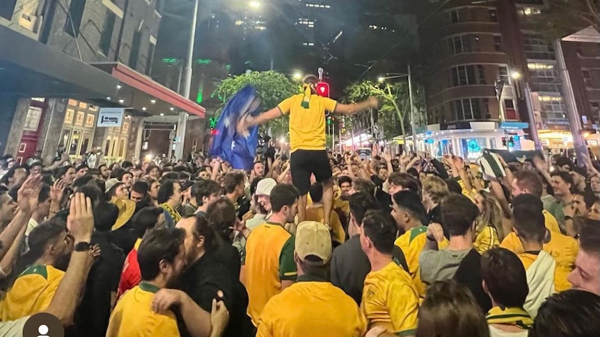 A crowd of people wearing Australian soccer shirts stand on a city street and raise their hands in triumph.