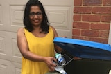 A woman in a yellow top smiles at the camera as she puts some recycling into a bin outside her home.