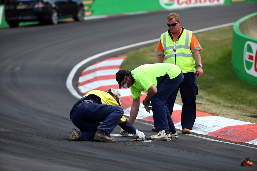 Workmen crouch and kneel on the tarmac on a race track trying to smooth the cracked suface during a race at Bathurst.