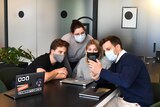 Four workers sit around a table wearing face masks and looking at a phone, in a story about managing back-to-work anxiety.