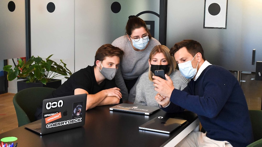 Four workers sit around a table wearing face masks and looking at a phone, in a story about managing back-to-work anxiety.