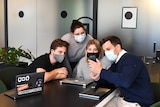 Four workers sit around a table wearing face masks and looking at a phone, in a story about managing back-to-work anxiety.