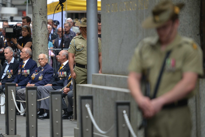 Elderly veterans wearing medals sit while Australian soldiers stand in front of them at a Cenotaph in Sydney.