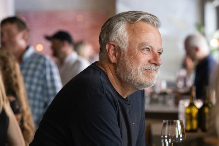 Side shot of a middle-aged man with grey hair, grey beard and a dark blue shirt sitting in a restaurant with a glass of wine. 