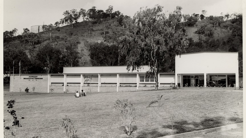 A low and long building set against a hill. A car parked at the entrance and two people sit in the foreground.