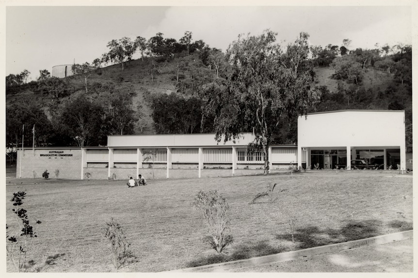 A low and long building set against a hill. A car parked at the entrance and two people sit in the foreground.