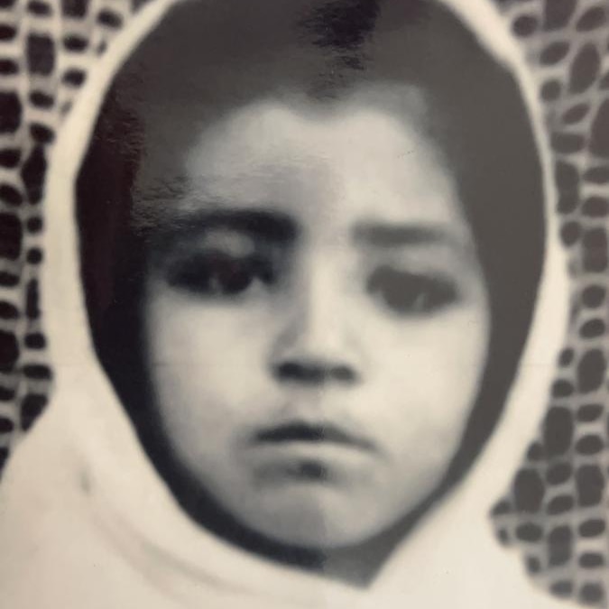 A little girl wearing a headscarf looks straight into the camera in a black and white image.
