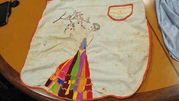 An old and faded white linen apron with embroidery of a woman in a colourful dress reaching for flowers.