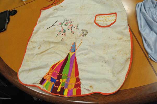 An old and faded white linen apron with embroidery of a woman in a colourful dress reaching for flowers.