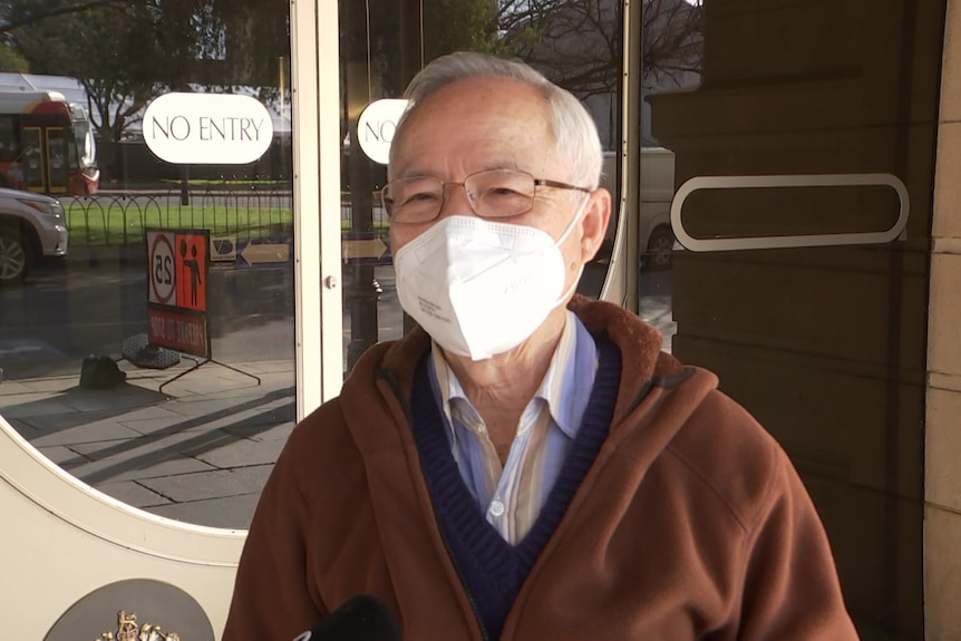 A man with short grey hair and glasses with a face mask on stands in front of a court building