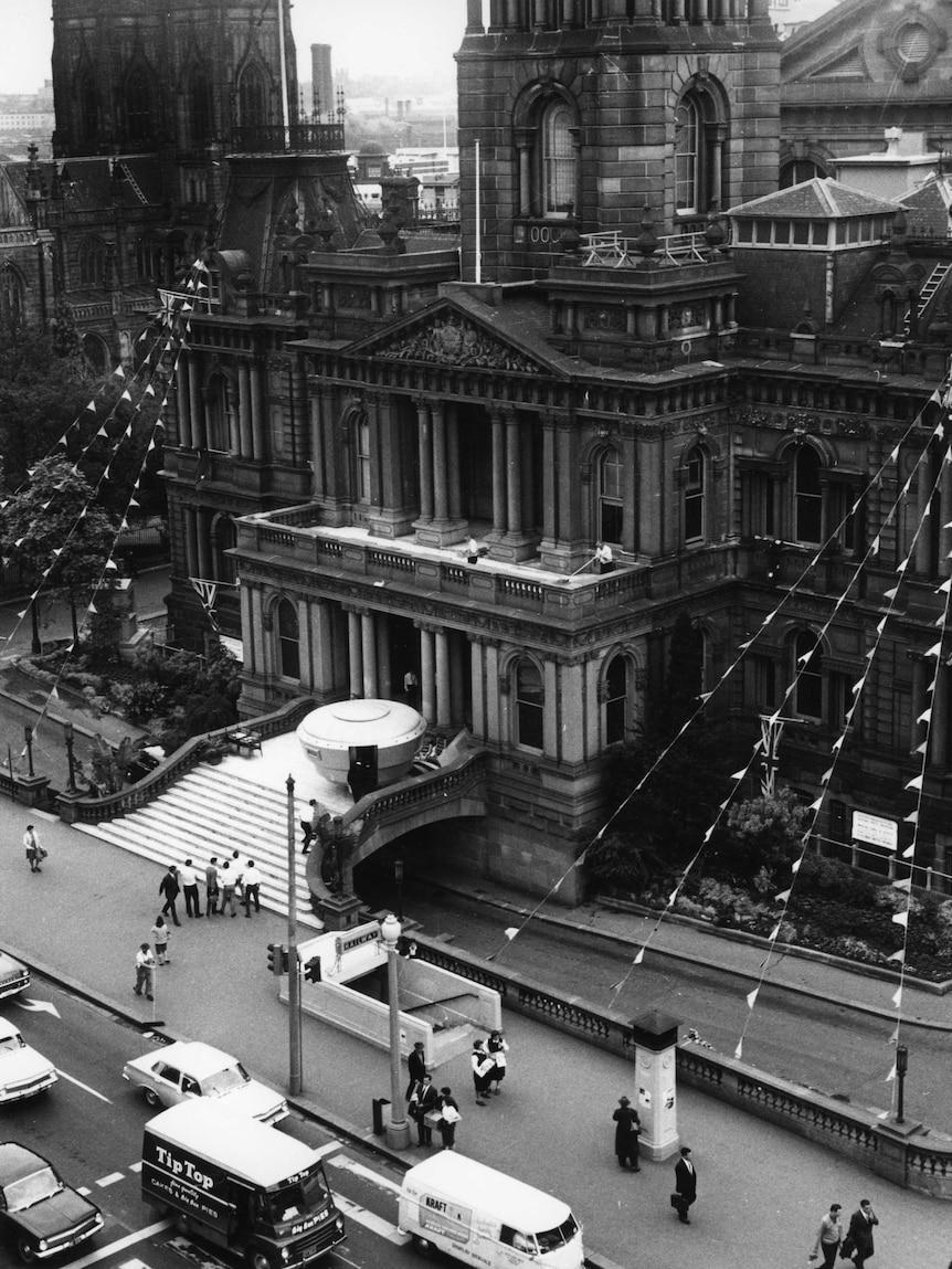 Black and white photo of UFO model on steps of Town Hall with people and cars in street in 1965.