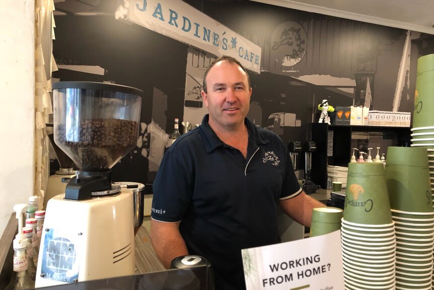 A man in a navy polo smiles at the camera behind a coffee machine with a sign saying "Jardine's Cafe" behind him.