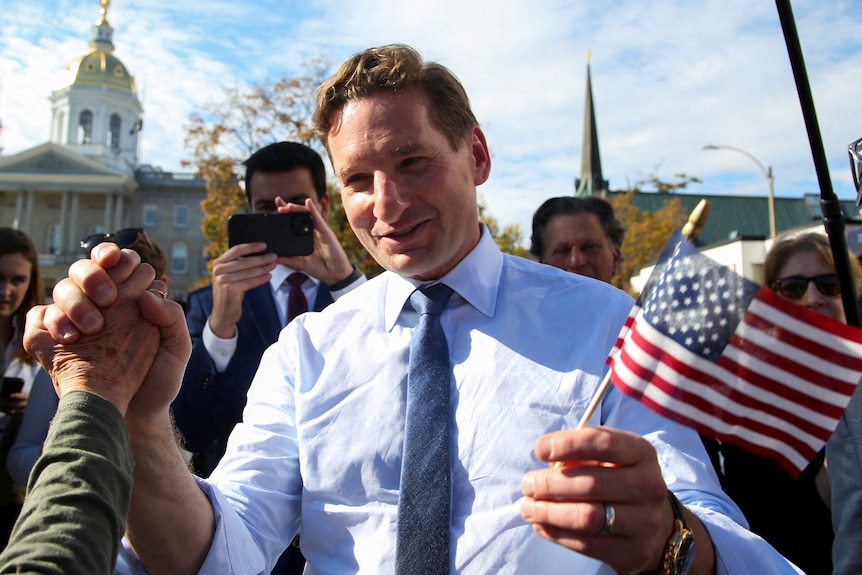 A middle-aged man in a light blue business shirt smiles and holds a small American flag as he grasps hands with another person.