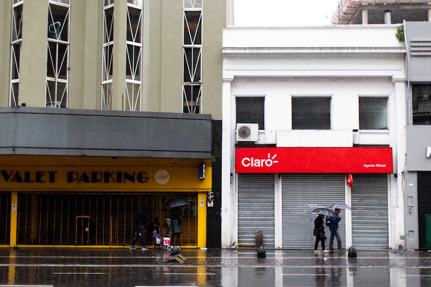 A couple holding umbrellas walk past a row of closed and dark shops