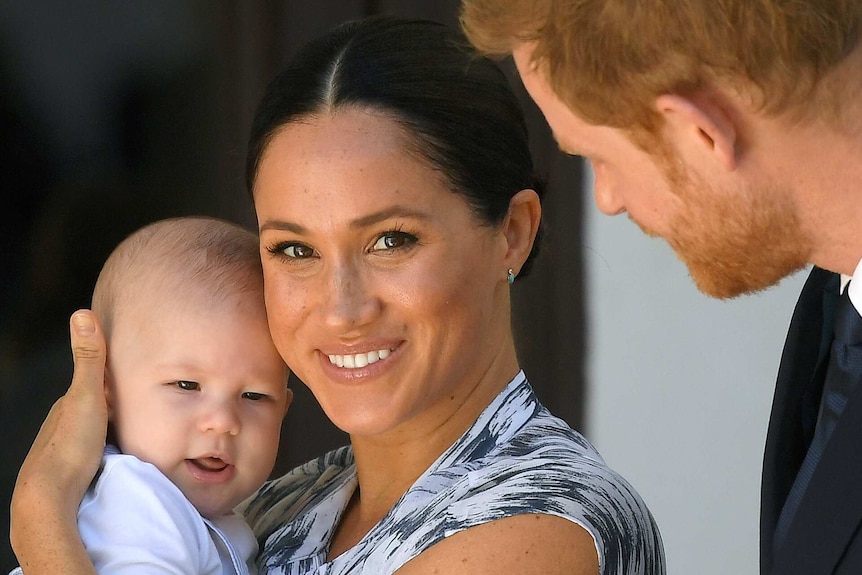 Meghan looks at the camera and smiles while holding Baby Archie. Prince Harry leans towards them, but is facing away from shot.