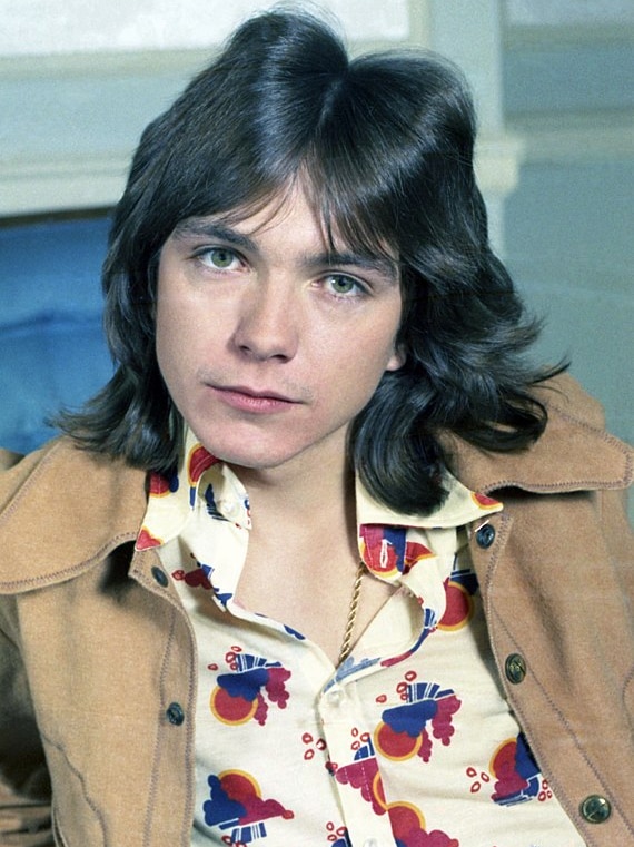 David Cassidy leans back and looks into the camera, wearing a brown jacket and a button-up shirt with a colourful print