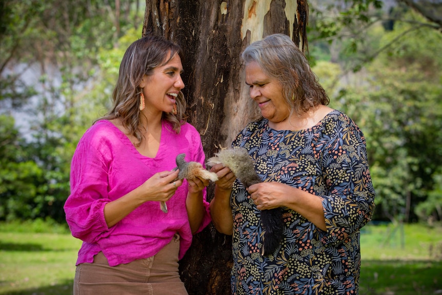 Kirli Saunders and Walbunja elder Aunty Loretta smiling and talking in bushland outdoors, while holding possum parts.