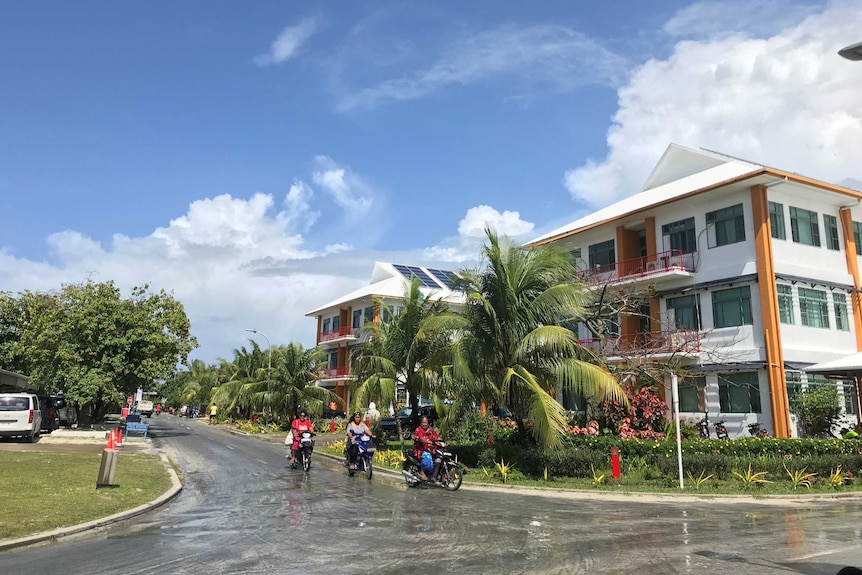 People ride motorcycles down a tree lined suburban street.